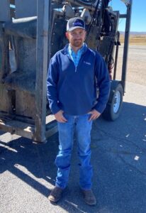 Dr. Sam is smiling as he stands beside his equipment, dressed in a blue jacket, jeans, and a cap, looking ready for the day's work.