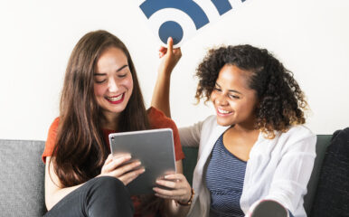 Two women sitting on a sofa, smiling and using a tablet together. One of them is holding a large Wi-Fi signal icon above their heads, symbolizing a strong internet connection.