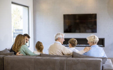 A multi-generational family sitting together on a sofa in a modern living room, enjoying quality time while watching free cable TV. The group includes grandparents, parents, and children, all facing the television, creating a warm and cozy atmosphere.