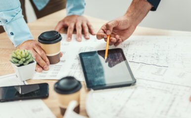 Two professionals leaning over a desk, reviewing tablet plans displayed on a digital tablet while discussing architectural blueprints and documents, with coffee cups nearby.