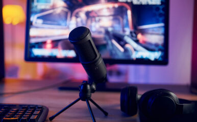 A high-quality microphone set up in front of a computer monitor displaying a shooter game, ready for supreme streaming of gaming content with headphones nearby.