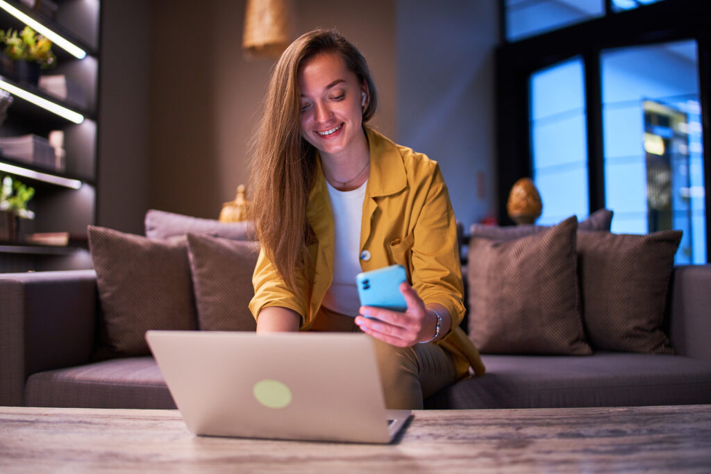 A young woman smiling while using her smartphone and laptop simultaneously in a modern, cozy room, illustrating the efficiency of multitasking with a high-speed internet connection and considering if 1000 Mbps is fast.