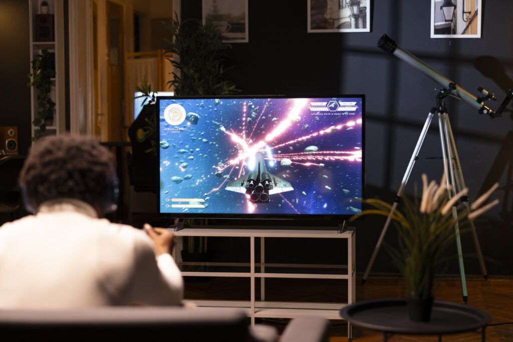 A person sitting in a dark room with a telescope in the background, playing a space-themed video game on a TV screen. This image illustrates "how to stream TV without cable," showcasing alternative entertainment options like gaming through streaming platforms.