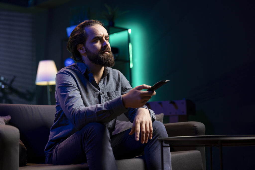 A man sitting on a sofa in a dimly lit room, holding a remote control, engaged in supreme streaming while selecting shows or movies to watch.