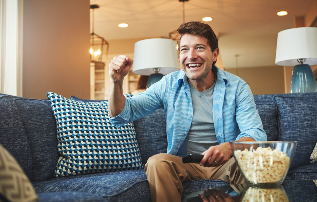 A man sitting on a sofa with a bowl of popcorn, cheering while watching a sports game on his TV, symbolizing excitement and entertainment choices between Roku TV and Google TV.