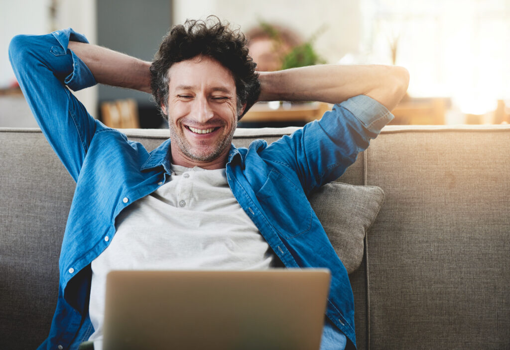 A happy man relaxing on his couch at home, smiling while looking at his laptop screen. This image represents the ease and comfort of accessing a fast home fibre optic connection for uninterrupted browsing or work from home.