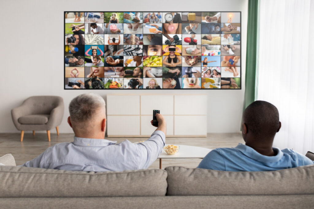 Two men sitting on a couch in a living room, comparing and watching different streaming content on a large television screen, illustrating a decision between Roku TV and Google TV.