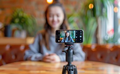 A young woman sitting at a table, recording herself with a smartphone on a tripod, as she creates content, possibly sharing behind-the-scenes details on how it's made streaming from a cozy café environment.