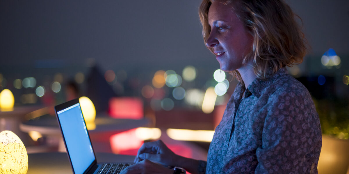 A woman working on a laptop at a rooftop bar during nighttime, with colorful lights in the background, representing the high-speed internet required for remote work and answering the question: is 1000 Mbps fast?