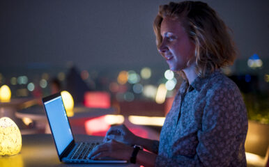 A woman working on a laptop at a rooftop bar during nighttime, with colorful lights in the background, representing the high-speed internet required for remote work and answering the question: is 1000 Mbps fast?