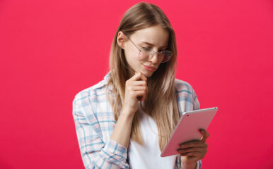 A young woman with glasses, holding a tablet and thinking against a pink background, possibly considering how to test her ping for an online task.