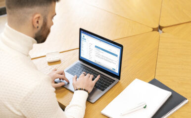 A young man in a white sweater typing on a laptop in a bright, modern study environment, reflecting a focused approach to completing tasks related to the "Internet Essentials application.