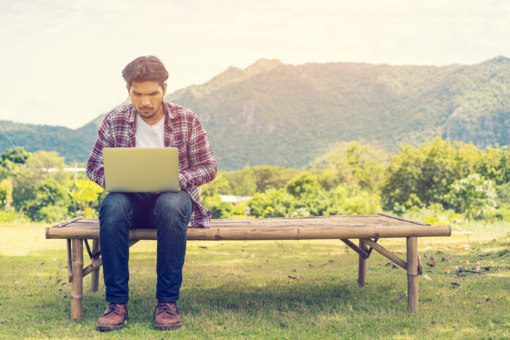  A young man sits on a wooden bench outdoors, using a laptop with a focused expression. He is surrounded by a lush green landscape with mountains in the background, depicting the challenge of getting reliable internet in remote areas. Keywords: how to get internet in rural areas.