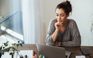 A young woman in a home office environment, gazing intently at her laptop screen with a pen in hand, reflecting on her search for an "internet download manager crack" to manage her digital downloads more efficiently