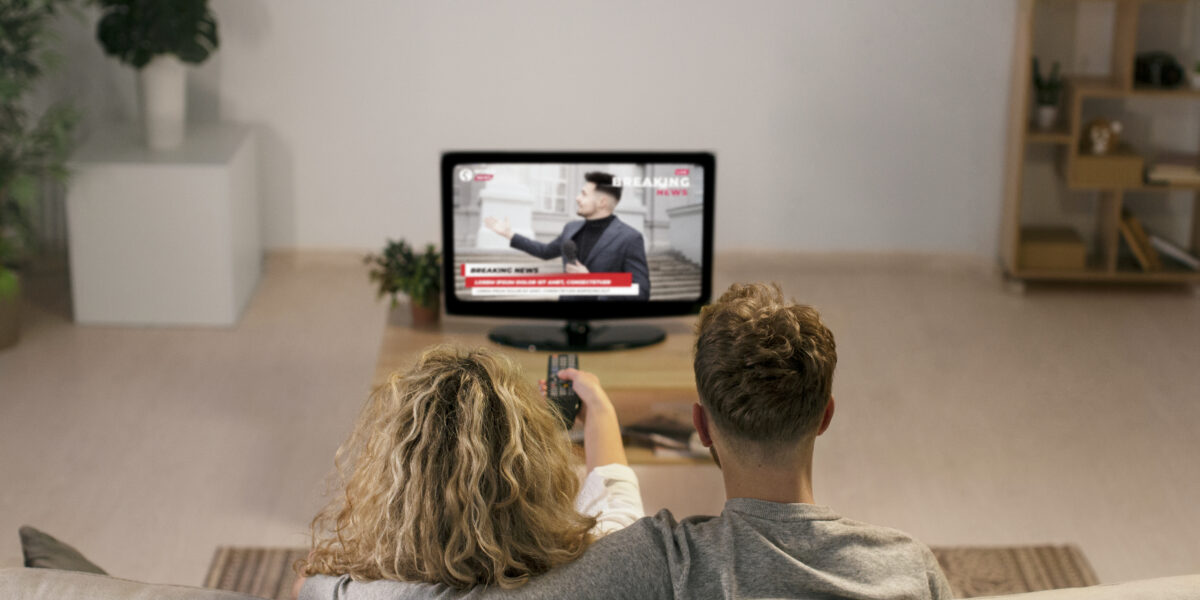 Back view of a couple sitting on a sofa, watching breaking news on Google TV. The cozy setting highlights the home streaming experience, possibly from YouTube’s news channel.