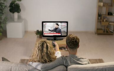 Back view of a couple sitting on a sofa, watching breaking news on Google TV. The cozy setting highlights the home streaming experience, possibly from YouTube’s news channel.