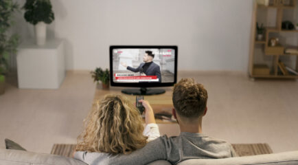 Back view of a couple sitting on a sofa, watching breaking news on Google TV. The cozy setting highlights the home streaming experience, possibly from YouTube’s news channel.