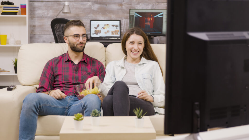 A young couple sitting on a couch, enjoying snacks while watching a show on Google TV, engaged and smiling. YouTube videos are open on the computer in the background.