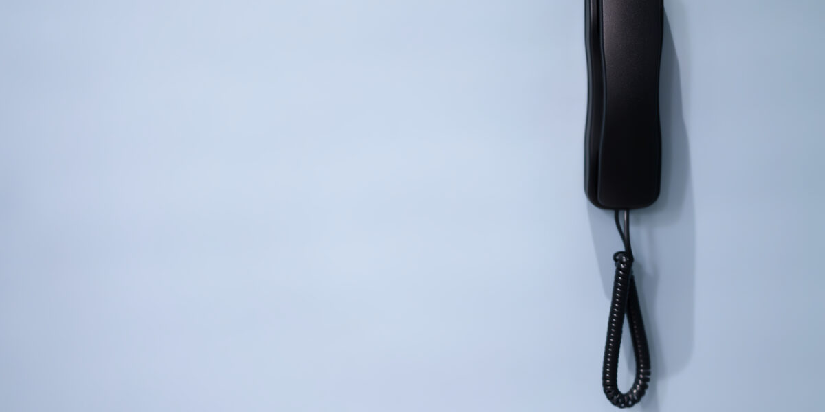 A black basic phone handset hanging against a light blue wall, creating a minimalist visual with ample negative space.