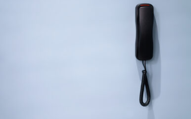 A black basic phone handset hanging against a light blue wall, creating a minimalist visual with ample negative space.