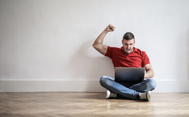 A cheerful man celebrates a successful internet connection while sitting on the floor with his laptop, symbolizing excitement after using a test my speed service to confirm fast performance.