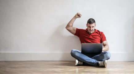 A cheerful man celebrates a successful internet connection while sitting on the floor with his laptop, symbolizing excitement after using a test my speed service to confirm fast performance.