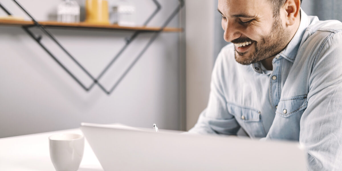 A smiling entrepreneur working in a cozy home office, engaged with his laptop while sipping coffee, showcasing a relaxed setup perfect for checking internet speed. Keywords: test my internet speed.