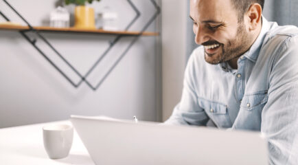 A smiling entrepreneur working in a cozy home office, engaged with his laptop while sipping coffee, showcasing a relaxed setup perfect for checking internet speed. Keywords: test my internet speed.