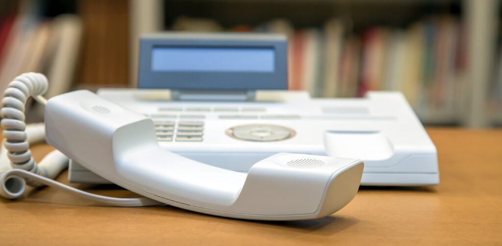 A close-up of a white basic phone with the handset resting on a desk, showing the keypad and display, ideal for showcasing classic office communication equipment.