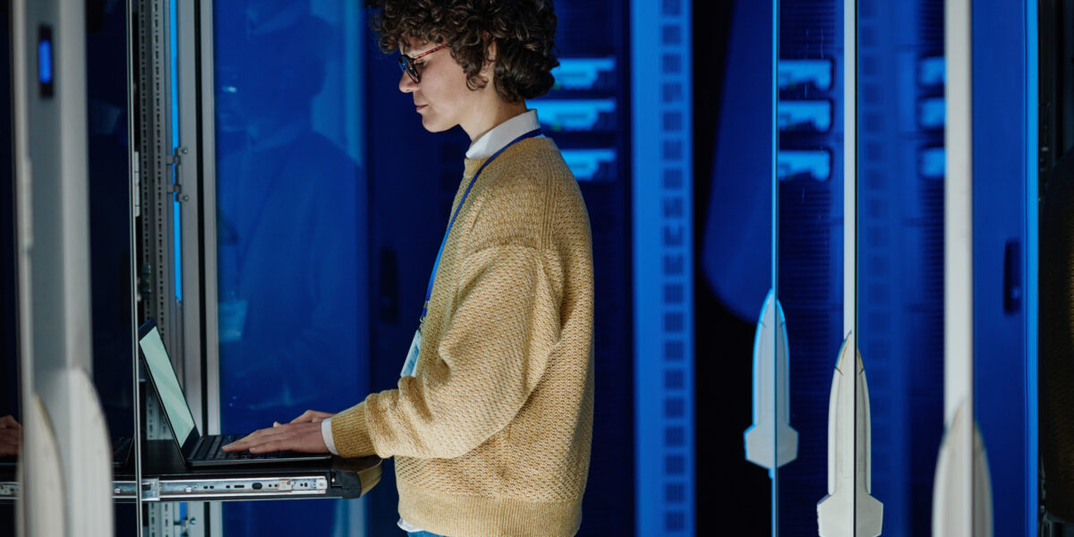 A technician wearing a beige sweater and glasses works on a laptop in a server room with glowing blue panels. This image represents "residential ISP solutions" with a focus on technical infrastructure and troubleshooting.