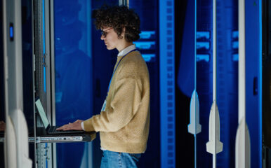 A technician wearing a beige sweater and glasses works on a laptop in a server room with glowing blue panels. This image represents "residential ISP solutions" with a focus on technical infrastructure and troubleshooting.