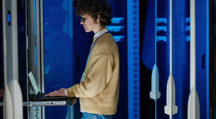 A technician wearing a beige sweater and glasses works on a laptop in a server room with glowing blue panels. This image represents 