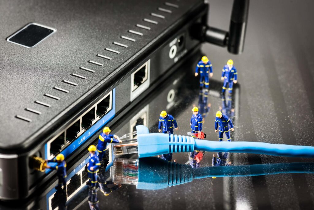 Creative representation of a broadband network provider's technical work. Miniature figures in blue uniforms appear to "work" on a blue Ethernet cable and router ports, symbolizing network connection and troubleshooting.