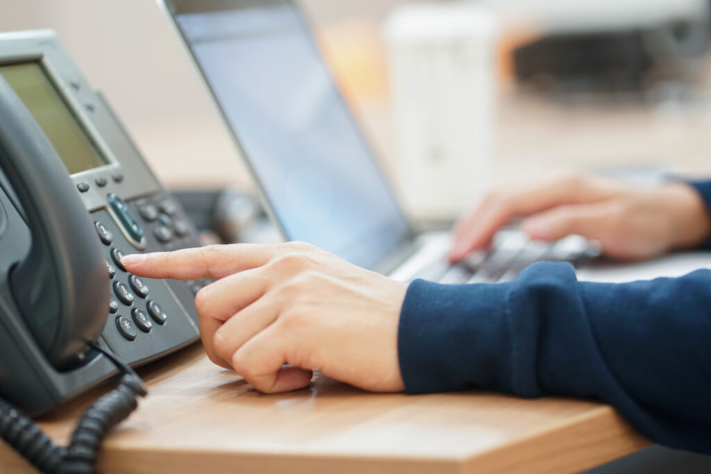 A person uses a landline phone while working on a laptop at their desk, demonstrating the efficiency of the best landline phone service for professional and reliable communication.