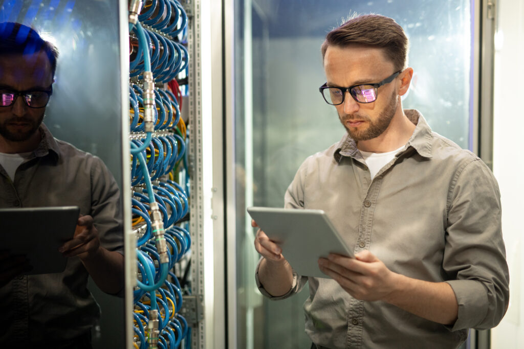 A broadband network provider engineer uses a tablet to inspect and monitor network connections in a data center. Rows of blue cables and server equipment surround the professional, emphasizing a high-tech working environment.