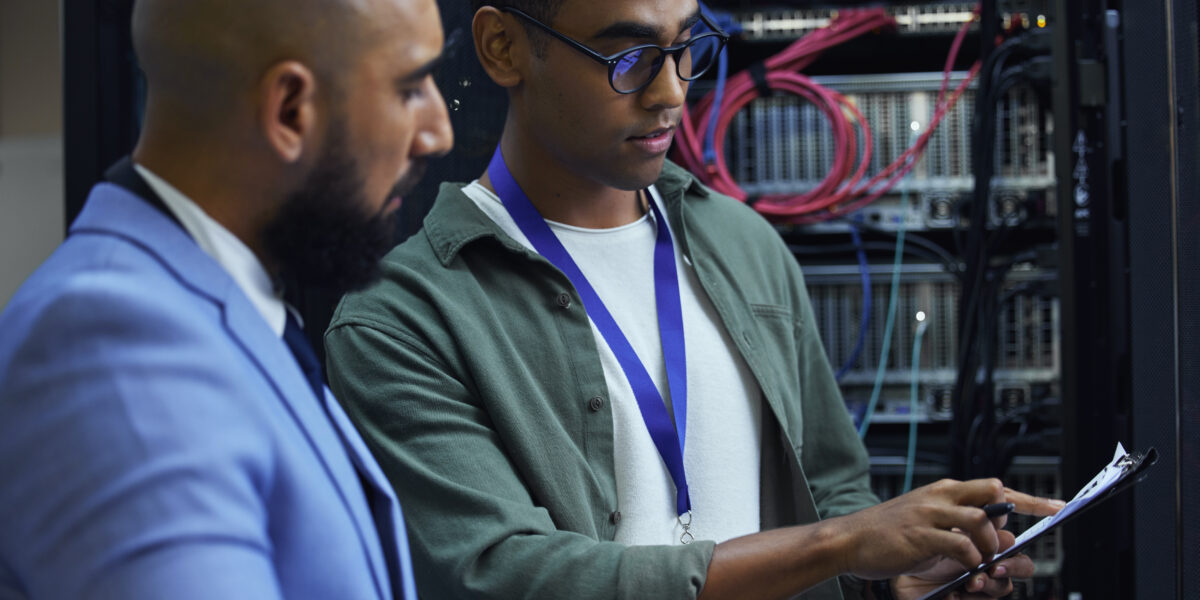 Two broadband network provider technicians are collaborating in a server room. One technician in a green shirt is pointing to a clipboard while the other, in a blue suit, attentively listens. The background shows server racks with organized network cables.