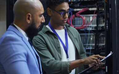 Two broadband network provider technicians are collaborating in a server room. One technician in a green shirt is pointing to a clipboard while the other, in a blue suit, attentively listens. The background shows server racks with organized network cables.