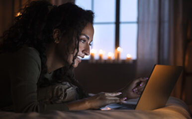 A woman smiling as she uses a laptop while lying on a bed lit by soft candlelight, representing the seamless connectivity provided by "internet fiber cable."