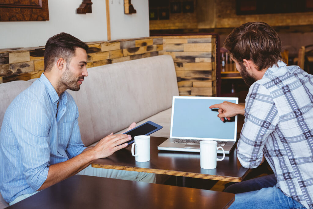 Two colleagues sit at a café, discussing work while using a laptop and tablet. This scene demonstrates the productivity benefits of the best cable internet deals, ensuring seamless online collaboration anywhere.