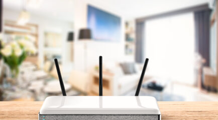 A sleek white WiFi router with three antennas is positioned on a wooden table in a brightly lit, modern living room. This image highlights the ideal central placement of the router for optimal connectivity in a stylish home setting. Keywords: WiFi router placement, modern home, best place to put WiFi router in house.