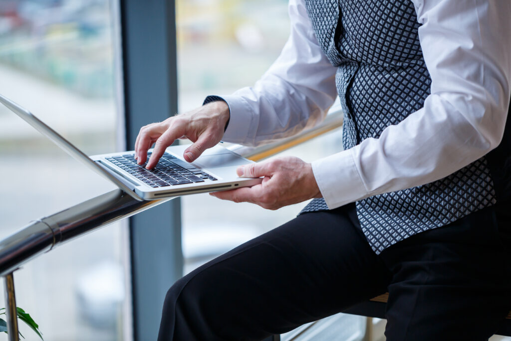 A businessman in a vest, sitting by a large window and typing on his laptop, emphasizing a sleek and productive workspace. Perfect for showcasing fiber for business benefits like high-speed internet for seamless operations.