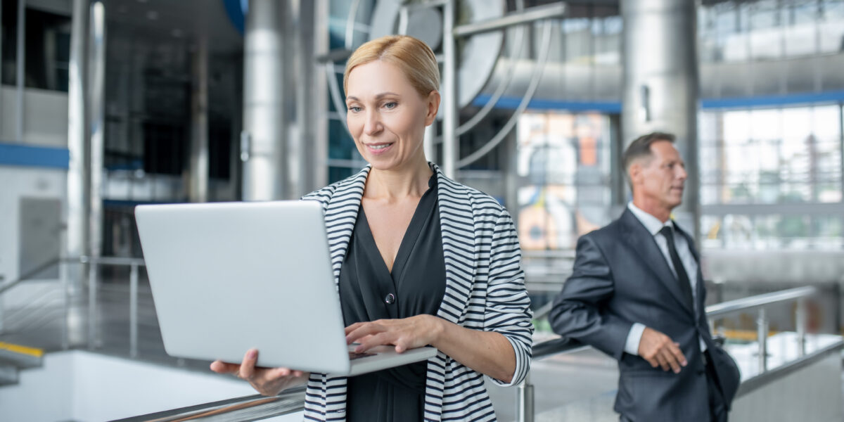 A professional woman smiling while working on her laptop in a modern, industrial-style building, with a business associate in the background. This is an excellent visual for promoting fiber for business solutions, highlighting reliability and professional connectivity.