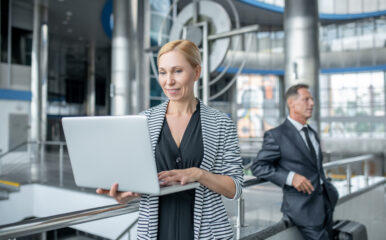 A professional woman smiling while working on her laptop in a modern, industrial-style building, with a business associate in the background. This is an excellent visual for promoting fiber for business solutions, highlighting reliability and professional connectivity.