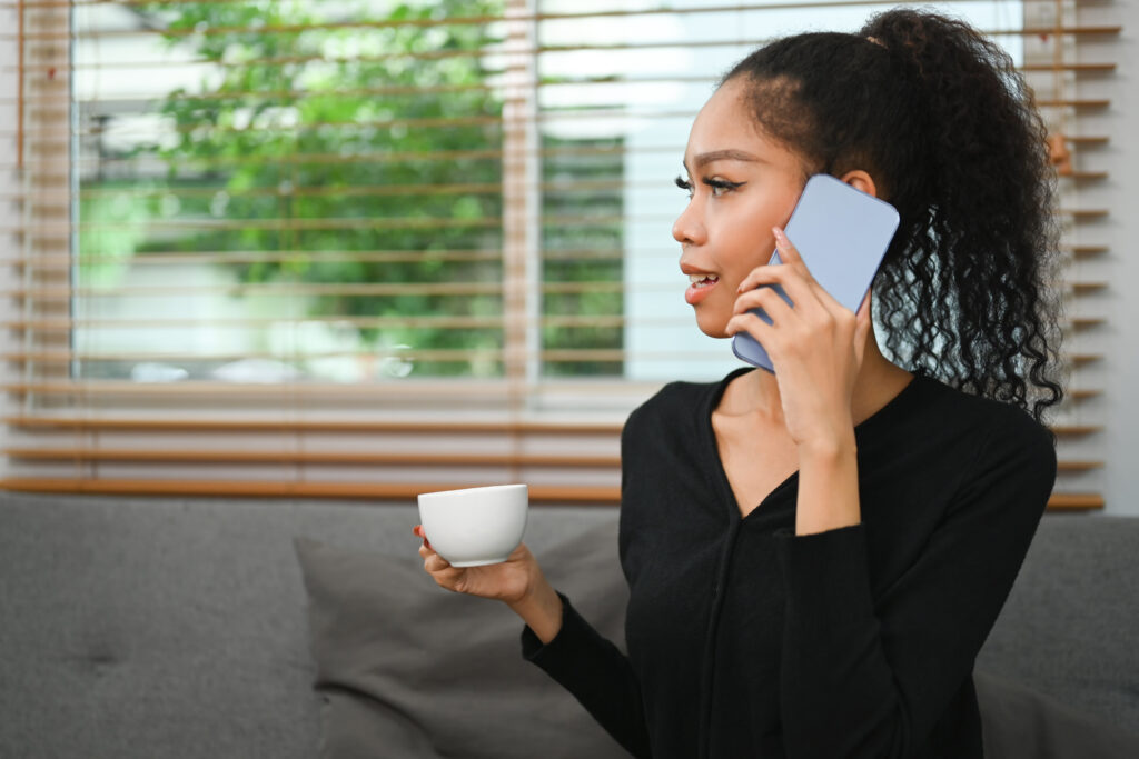  A young woman holding a coffee cup while talking on a smartphone, sitting in a cozy living room, highlighting "caller ID phone" features.