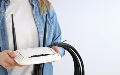 A person holding a white WiFi router with cables against a plain background, representing technical aspects and lifespan inquiries such as "how long do WiFi routers last."