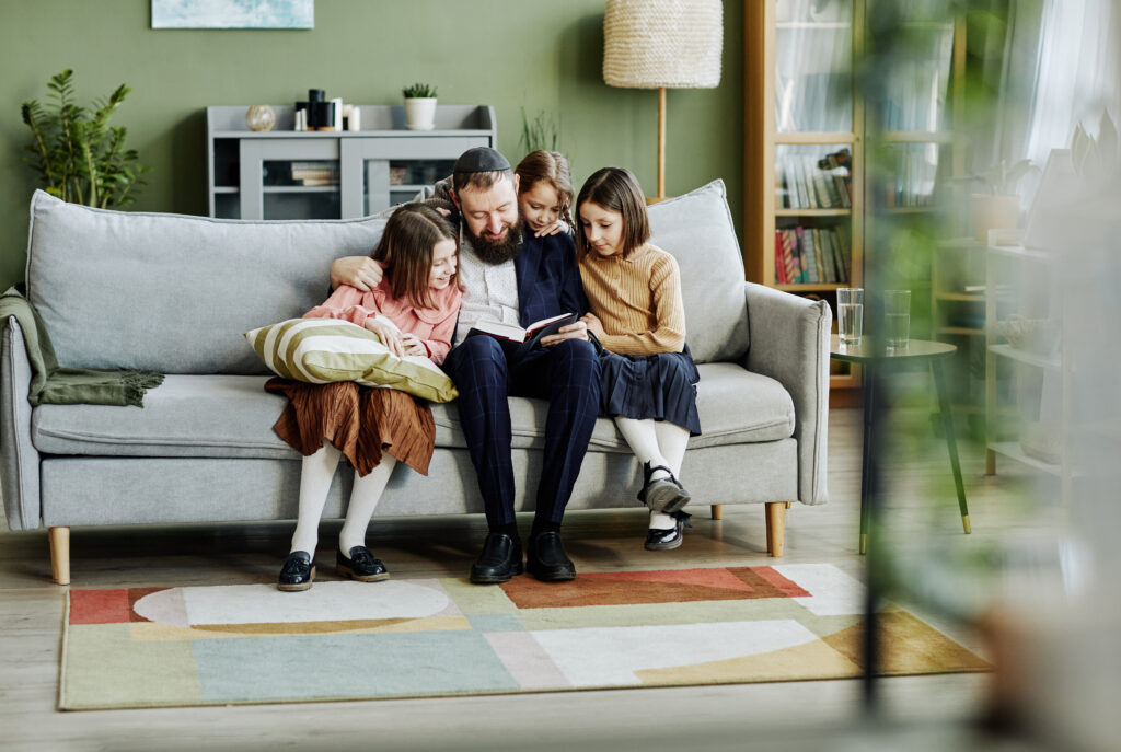 A warm family moment with an Orthodox Jewish father reading to his three children on a gray couch in a modern living room, representing family connectivity. Keywords: "best place to put wifi router in a 2 story house."