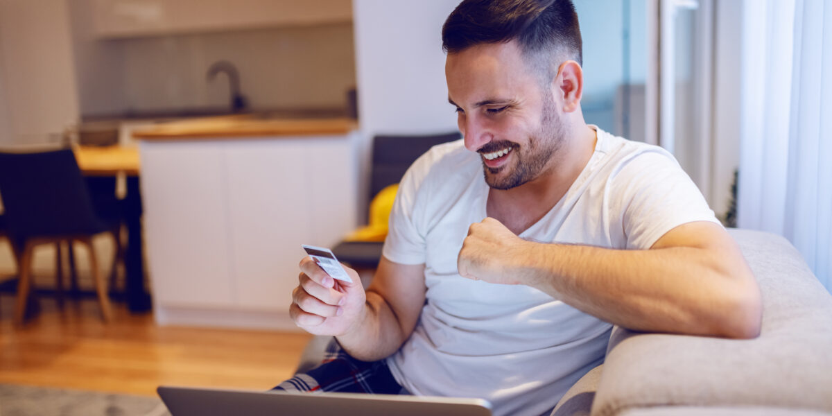 A smiling man sitting on a sofa, using a laptop and a credit card for online shopping, showcasing the convenience of high-speed internet connectivity at home.