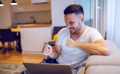 A smiling man sitting on a sofa, using a laptop and a credit card for online shopping, showcasing the convenience of high-speed internet connectivity at home.