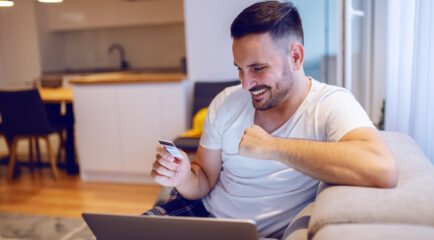 A smiling man sitting on a sofa, using a laptop and a credit card for online shopping, showcasing the convenience of high-speed internet connectivity at home.