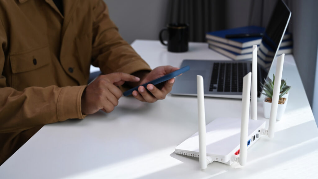  A person in a tan jacket sits at a desk, using a smartphone next to a white WiFi router with four antennas, surrounded by books and a laptop. The setup emphasizes the importance of placing the router near workspaces for uninterrupted internet access. Keywords: WiFi router placement, work desk, best place to put WiFi router in house.